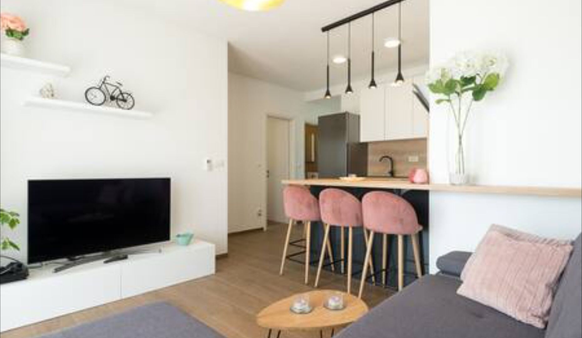 A modern living room with a flat-screen TV, a sofa, a coffee table, and a kitchen area with bar stools.
