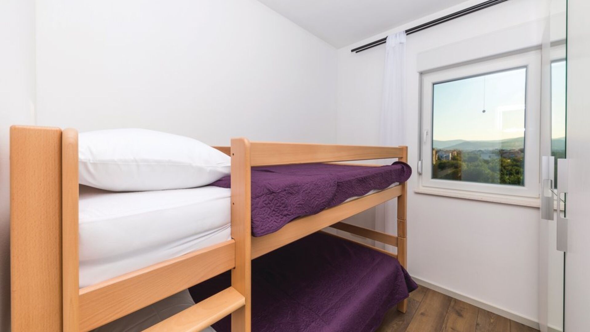 Een stapelbed met uitzicht uit het raam, witte wanden en een houten vloer.