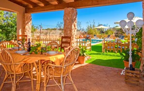 Une terrasse extérieure avec une table à manger pour deux personnes, entourée de chaises en osier, d’un pilier en pierre et offrant une vue sur une piscine et un jardin.