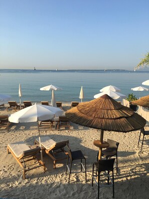 Een strand met rijen ligbedden en parasols, waarvan sommige voorzien zijn van rieten kappen, gericht op de zee.