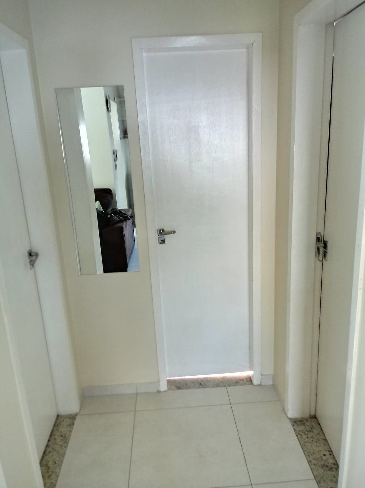 Un pasillo con una puerta blanca, un espejo en la pared izquierda y un piso de baldosas.