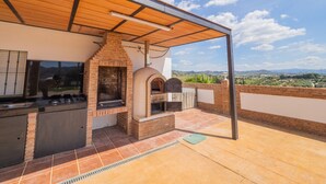Cocina exterior con horno de ladrillo y parrilla, bajo una pérgola de madera, con vistas a un paisaje pintoresco.