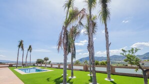 Zona de piscina con césped artificial y palmeras, rodeada por un muro de ladrillo y vistas a montañas distantes.