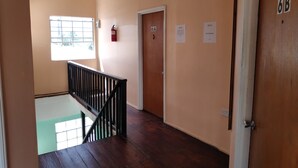 Un couloir avec un plancher en bois, une rambarde noire et une porte portant l’inscription « 6A ». Il y a une fenêtre sur la gauche et un extincteur accroché au mur.