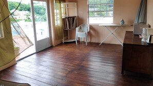 Une pièce avec un plancher en bois, une chaise blanche, une petite table et une fenêtre donnant sur un paysage verdoyant et des bâtiments.