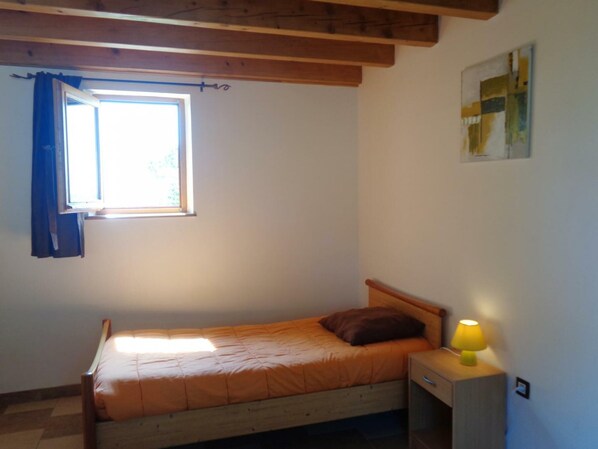 Une petite chambre avec un lit simple, une tête de lit en bois, une fenêtre avec des rideaux bleus et une table de chevet avec une lampe.