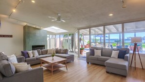 Ein geräumiges Wohnzimmer mit einer großzügigen Eckcouch, einem Couchtisch und einem Deckenventilator. Ein großes Fenster lässt natürliches Licht herein und bietet einen Ausblick ins Freie.