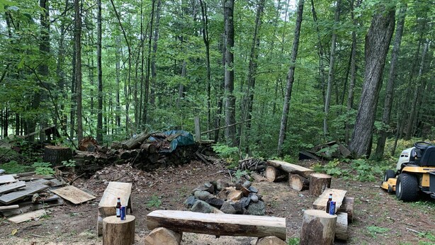Un claro en el bosque con bancos de madera y troncos, un tractor y leña dispersa.