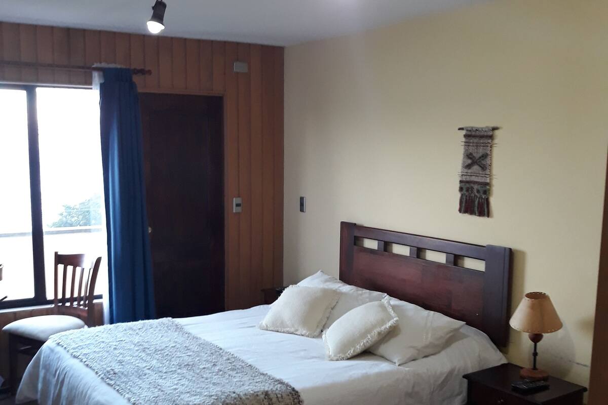 Une chambre à coucher avec un lit, une table de chevet, une lampe, une décoration murale et une fenêtre avec des rideaux.