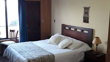 Um quarto com uma cama, mesa de cabeceira, luminária, um objeto pendurado na parede e uma janela com cortinas.
