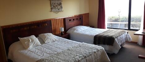 A hotel room with two beds, a wooden headboard, a nightstand, a lamp, a wall hanging, and a window with a view.