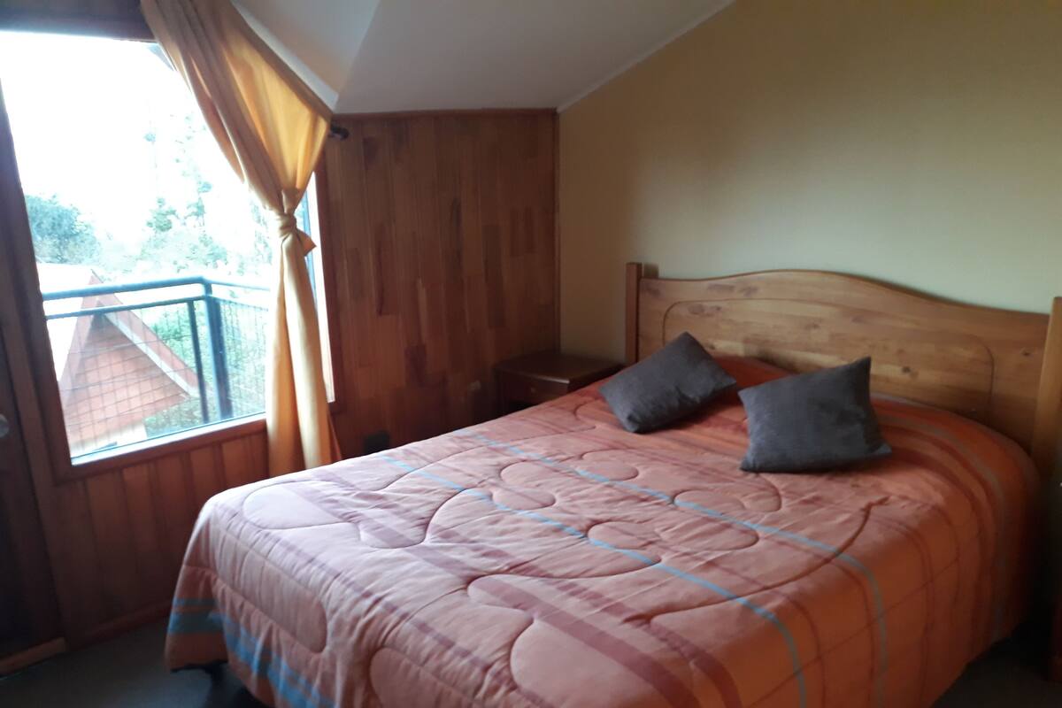 Une chambre à coucher avec une tête de lit en bois, une couvre-lit rose matelassée, deux oreillers, une table de chevet et une fenêtre avec des rideaux.