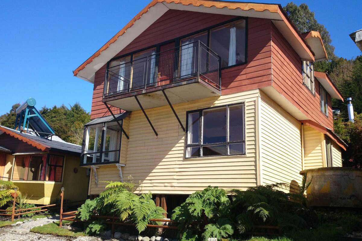 Une maison à deux étages avec un balcon, un petit jardin et un bâtiment bleu situé à côté.