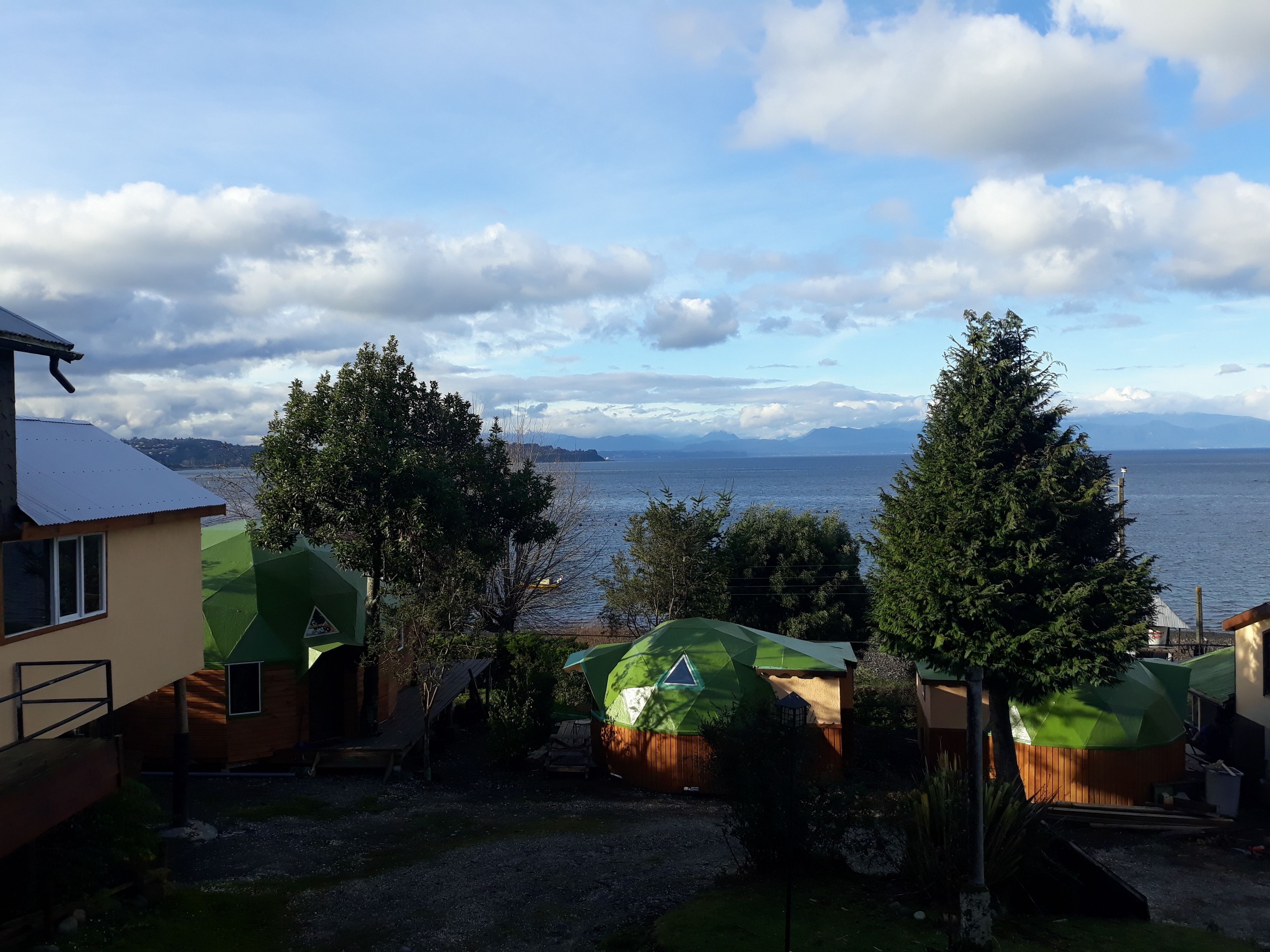 A coastal property with a view of the sea and mountains. There are several green-roofed structures, possibly cabins or houses, with a gravel driveway leading to them.