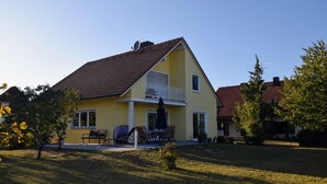Une maison jaune avec un toit rouge, un balcon et une terrasse aménagée avec du mobilier d’extérieur.