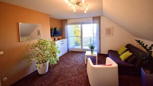 Un salon avec une grande fenêtre, un luminaire au plafond, un téléviseur à écran plat et une plante en pot.