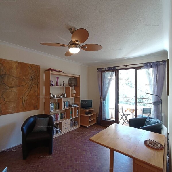 Un salon avec un ventilateur de plafond, une bibliothèque, un téléviseur, une table à manger et une vue par une fenêtre avec des rideaux.