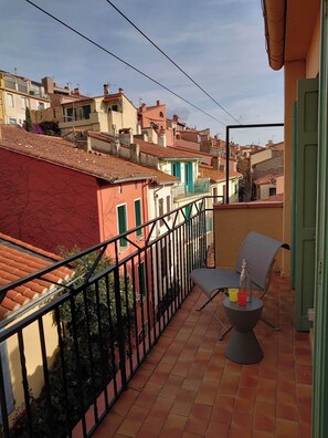 A balcony with a chair and table, overlooking a residential area with terracotta roofs and colorful buildings.