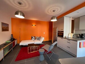 A compact room with a bed, a desk, a chair, a kitchenette, and a red rug.