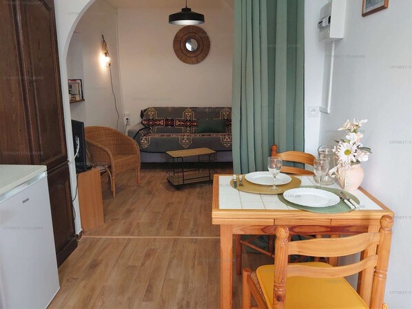 A dining area with a wooden table set for two, a sofa in the background, and a kitchen area with a refrigerator.