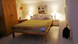 Un dormitori amb un llit de fusta, dues làmpades de nit, una tauleta de nit, un cistell de vimet i una prestatgeria penjada amb objectes decoratius.