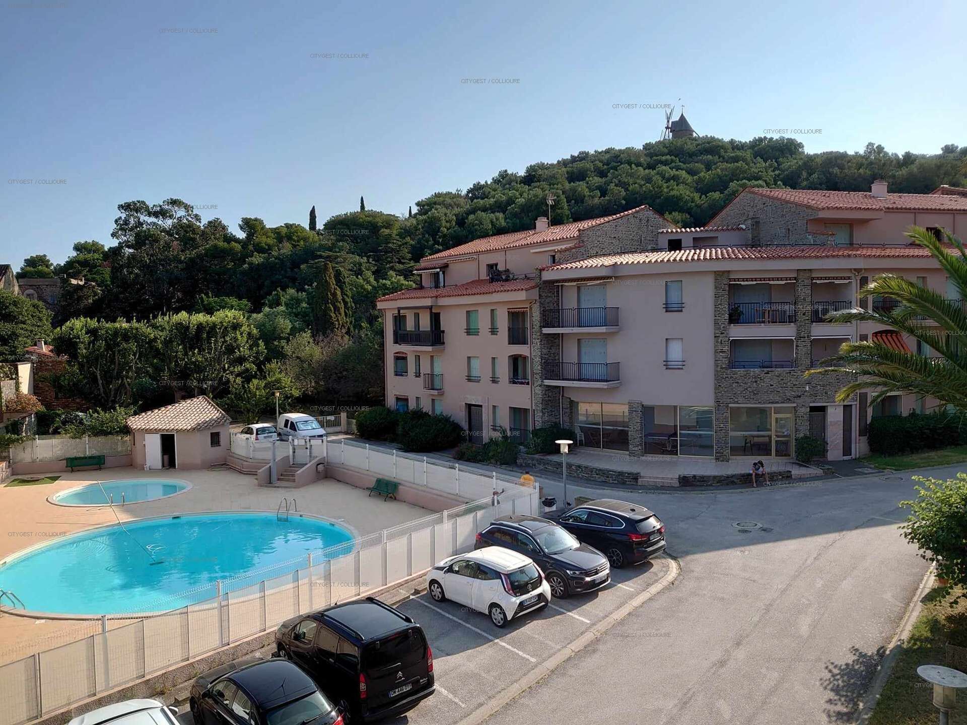 A multi-story residential building with a swimming pool, parked cars, and a hilly backdrop.