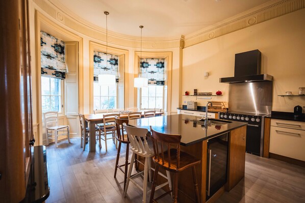 A kitchen with a central island, a stove, and a sink. There are chairs around the island and a dining table with chairs.