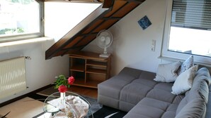 Een woonkamer met een bank, een glazen tafel met een bloemenvaas, een radiator, een ventilator en een raam met jaloezieën.