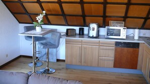 Een compacte keuken met een houten plafond, een kleine eettafel met een stoel, een magnetron, een broodrooster en een koffiezetapparaat.