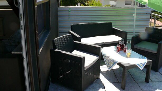 Een terras buiten met rieten meubels, een tafel met tafelkleed en een groene parasol.