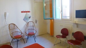 Ett rum med två fåtöljer av rotting med röda kuddar, en röd hopfällbar stol, en vit golvlampa, en stor spegel på väggen och en tv på ett stativ.
