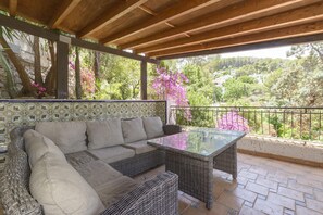 A covered patio with a wicker sofa and glass table, surrounded by a tiled floor and a railing.
