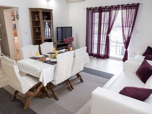 A dining area with a table set for breakfast, a television, and a view of the outdoors through purple curtains. - FAMILY  APARTMENT MLADEN AT NATURE PARK CLOSE TO THE SEA (Peracko Blato/Ploce)