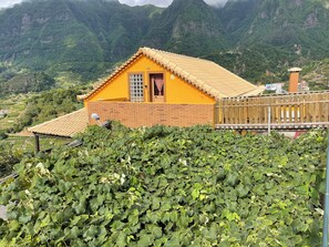 Een geel huis met een bakstenen muur en een pannendak, omringd door weelderige begroeiing en bergen.