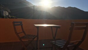 A terrace with two wooden chairs and a table, overlooking a mountainous landscape with the sun low in the sky. - Yellow Attic S. Vincent (São Vicente)