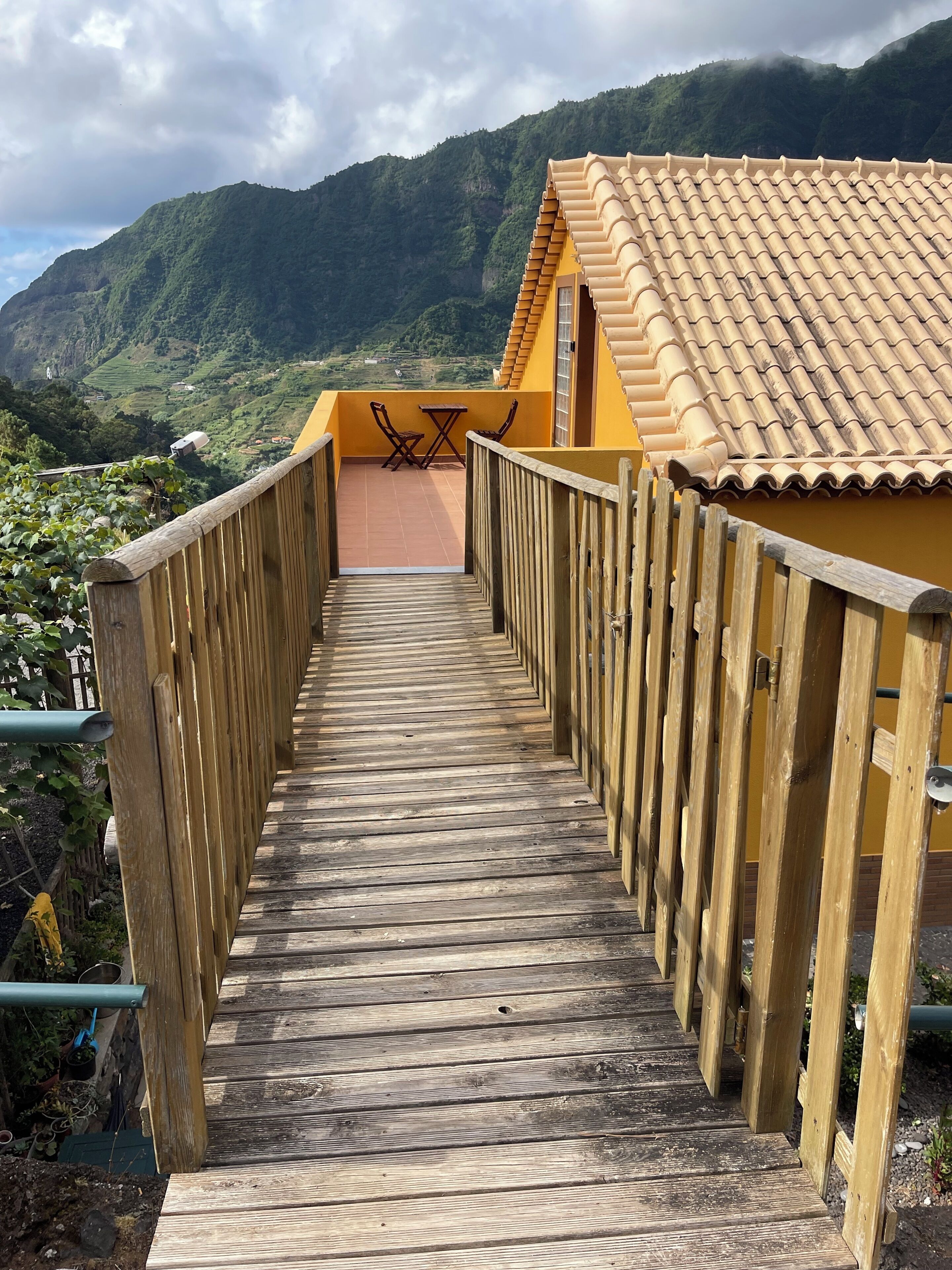 A wooden walkway leading to a yellow building with a tiled roof, surrounded by greenery and mountains.