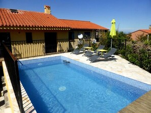 Una piscina con borde de baldosas, rodeada de sillones de descanso y sombrillas, y un edificio con tejado de tejas rojas.