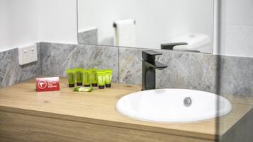 Un baño moderno con un tocador de madera, un lavamanos blanco y un espejo que refleja un inodoro. Hay artículos de aseo verde sobre el tocador.