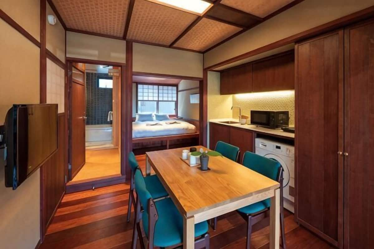 A compact room with a wooden dining table, chairs, a bed, a washing machine, and a microwave.