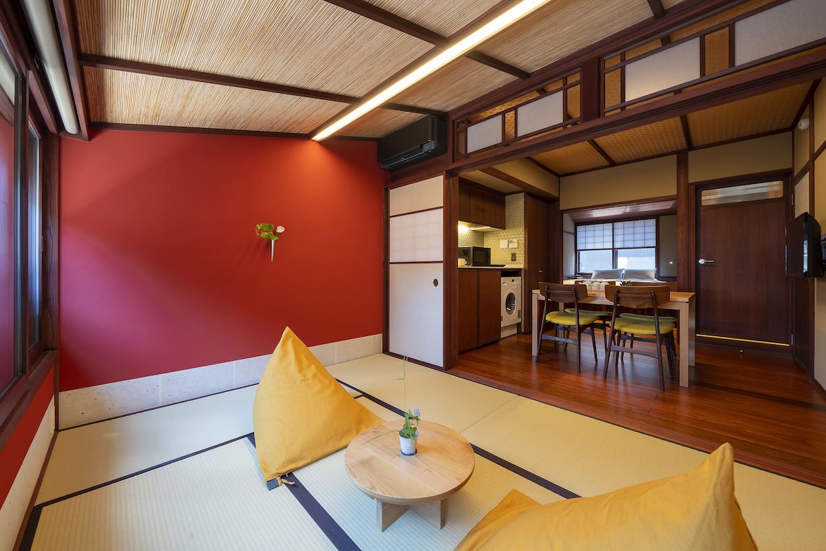 A traditional Japanese room with tatami flooring, a low wooden table with a plant, yellow cushions, and a kitchen area visible through a sliding door.