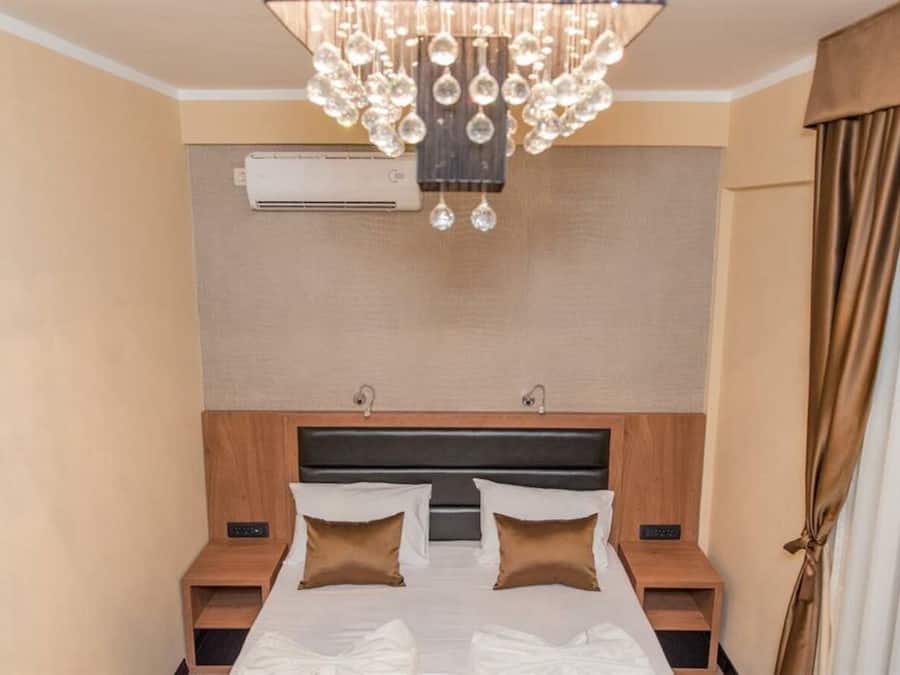A hotel room with a large bed, two bedside tables, a modern chandelier, an air conditioning unit, and a curtain on the window.