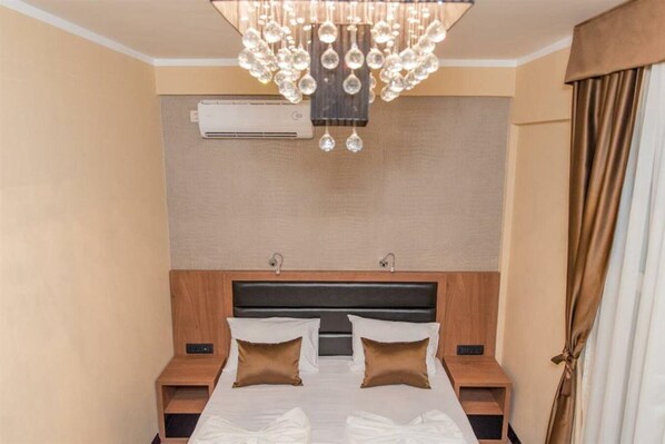 A hotel room with a large bed, two bedside tables, a modern chandelier, an air conditioning unit, and a curtain on the window. - Hotel Crnogorska Kuca (Podgorica)