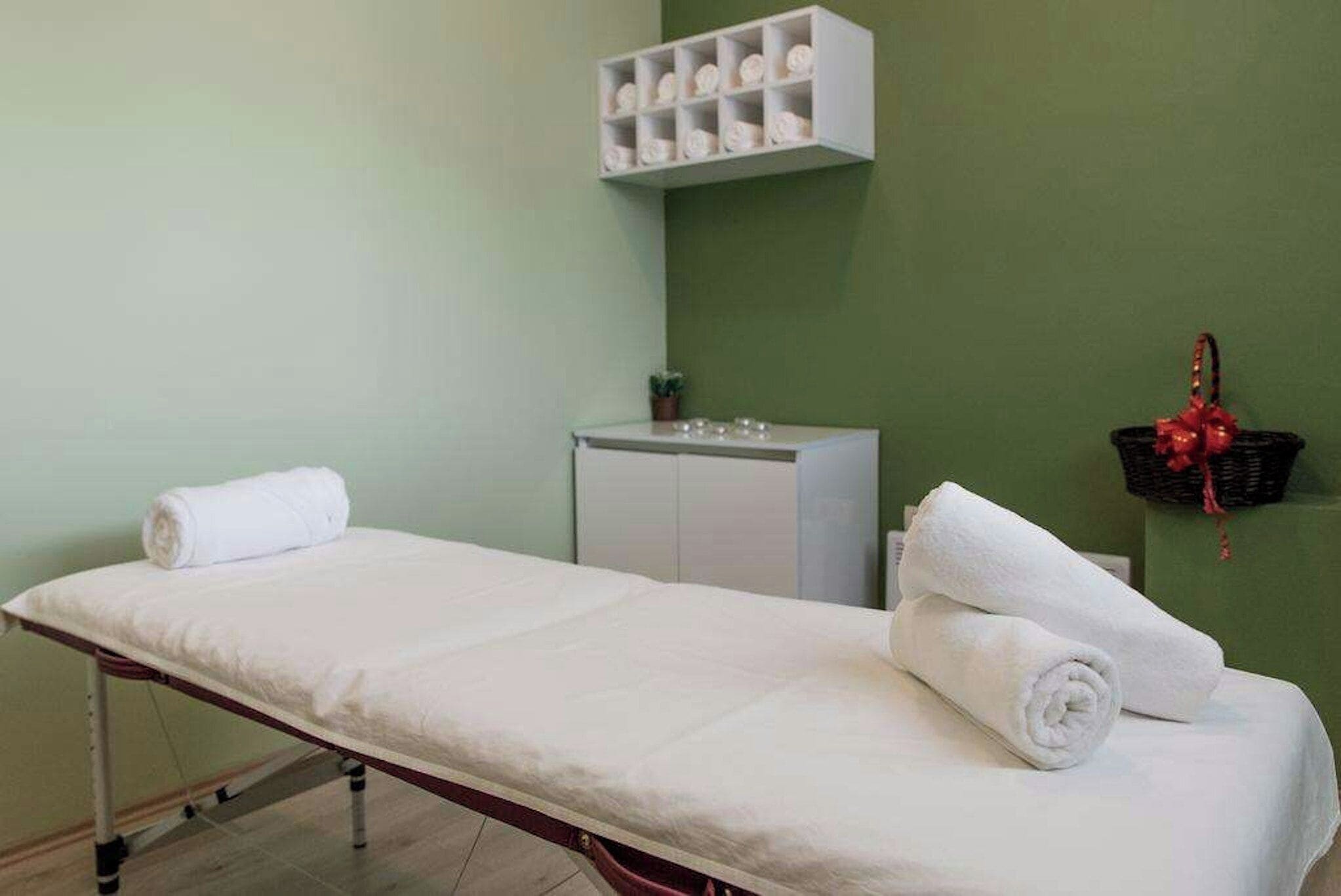 A clinical examination room with a single examination table, a white cabinet, and a wall-mounted shelf with rolled towels.