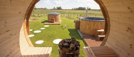 Una estructura de madera con una abertura circular, un brasero con la etiqueta 'HARVIA', y vistas a un área con césped, un jacuzzi y bancos de madera.