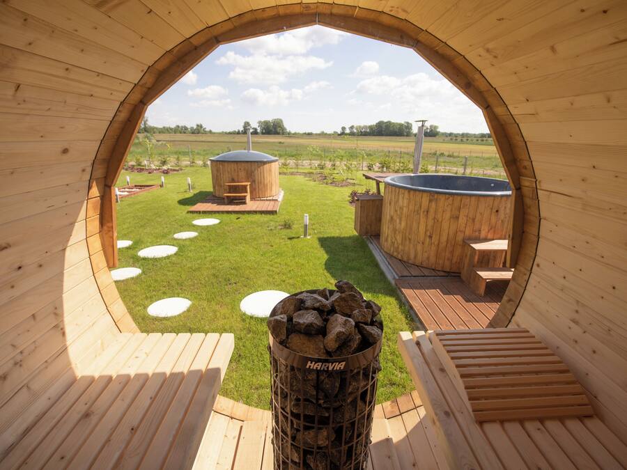 Una estructura de madera con una abertura circular, un brasero con la etiqueta 'HARVIA', y vistas a un área con césped, un jacuzzi y bancos de madera.