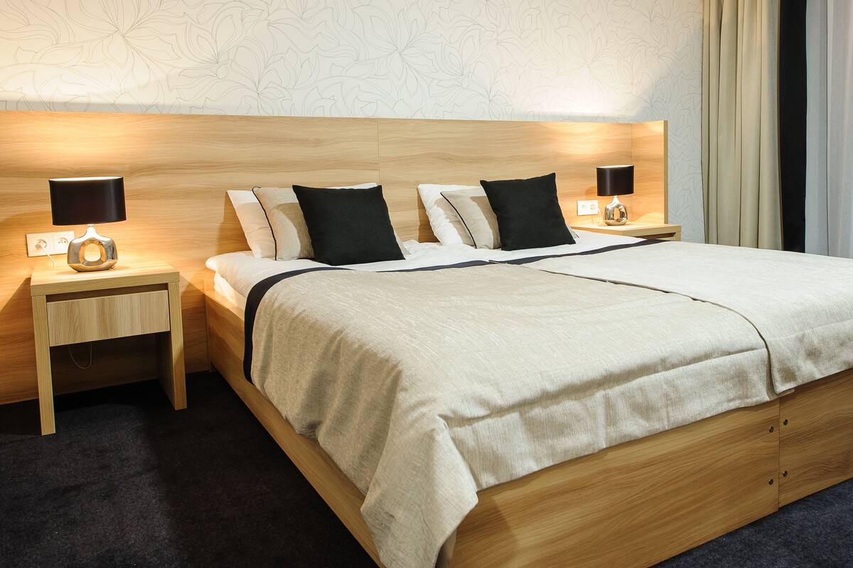 Una habitación de hotel con estructura de cama de madera, colcha color beige, dos almohadas negras, una mesita de noche con lámpara y un papel tapiz con motivos florales.