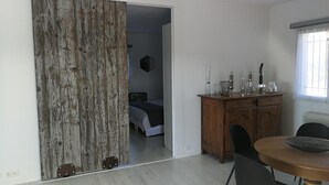 Quarto com guarda-roupa de madeira, cama com cabeceira preta, aparador de madeira com copos e mesa de jantar com uma cadeira.