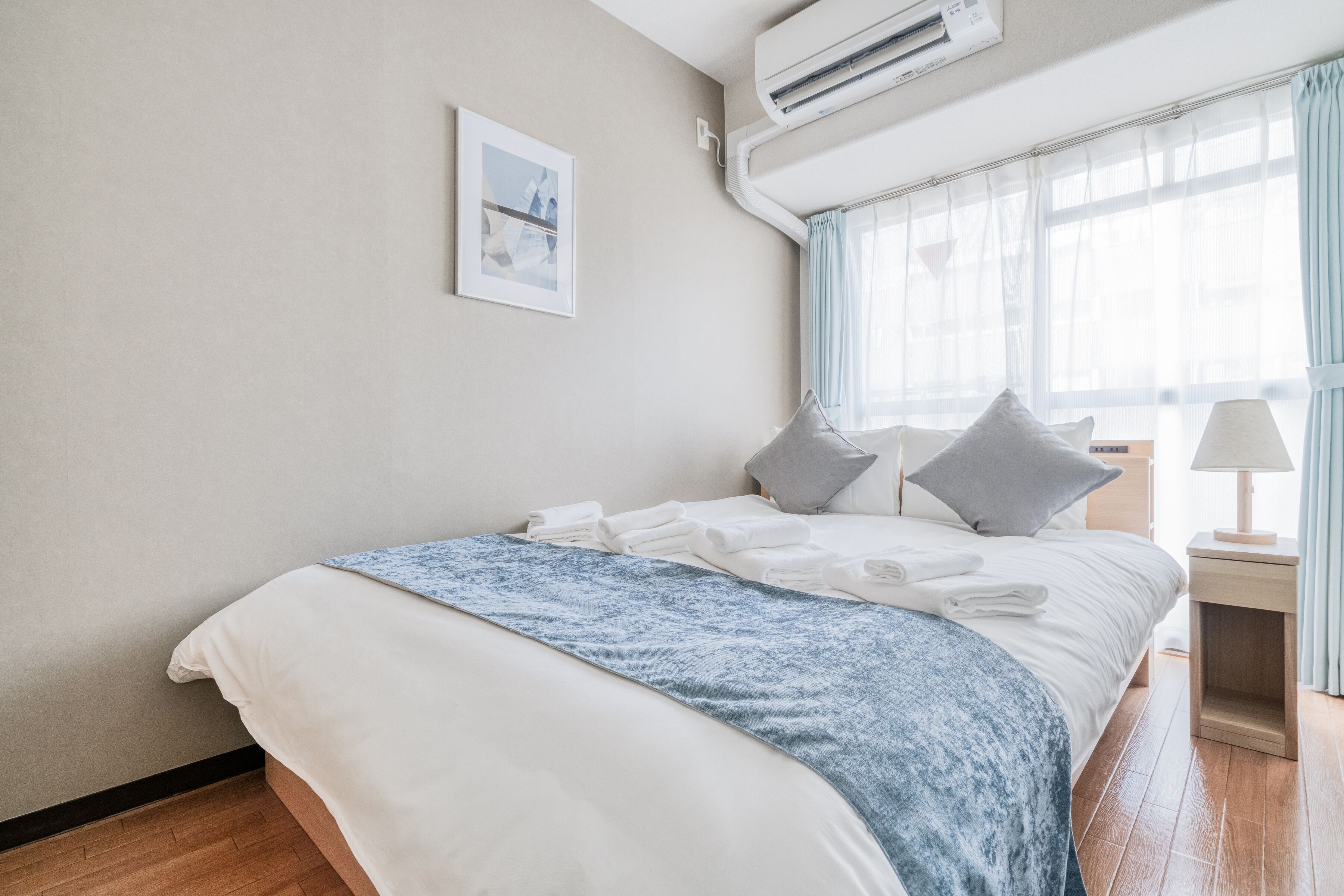 Una cama bien hecha con una manta azul, una mesita de noche con una lámpara y una ventana con cortinas traslúcidas.