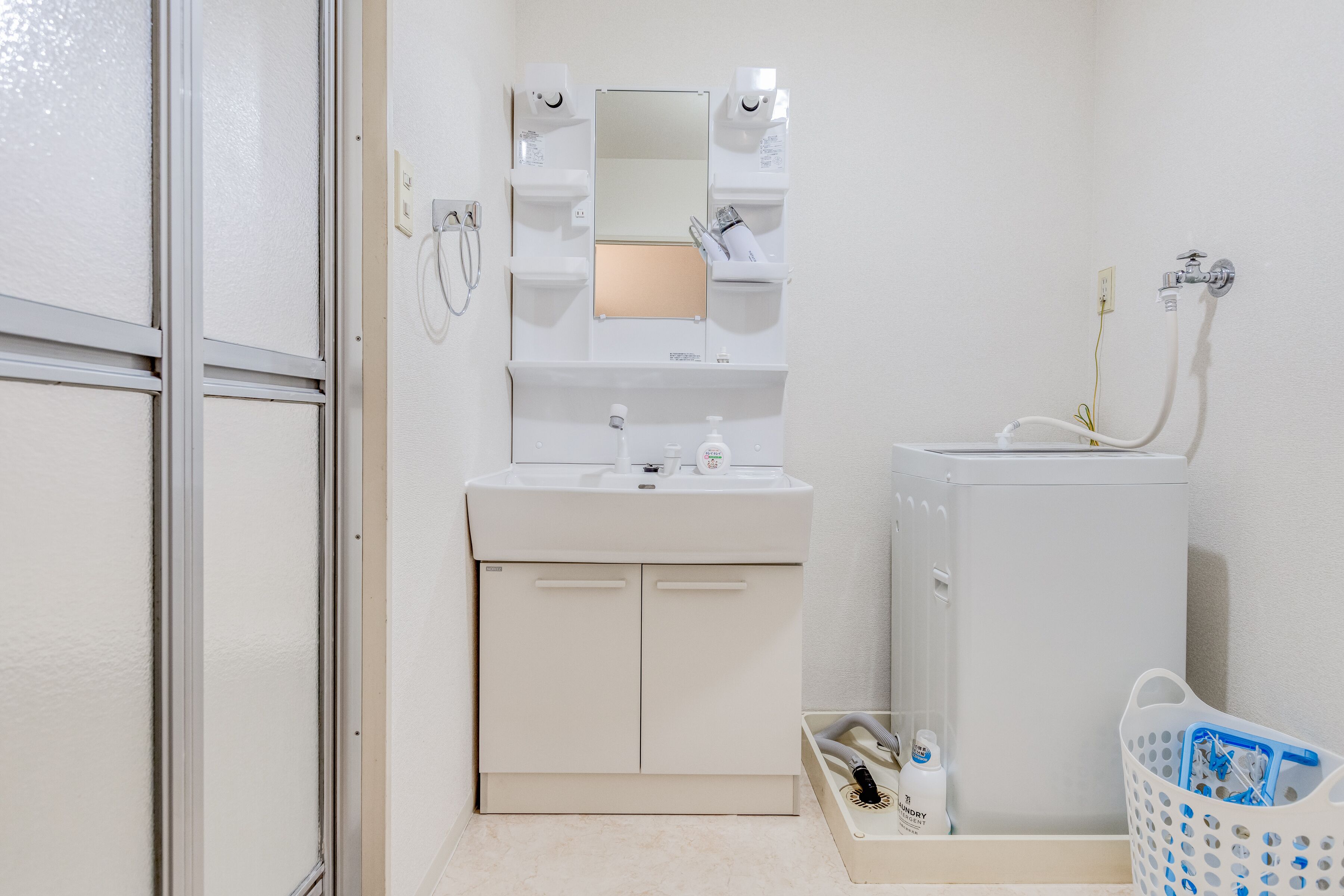 A small, white bathroom with a sink, mirror, and shower. There is a white laundry basket and a bottle of shampoo on the floor.