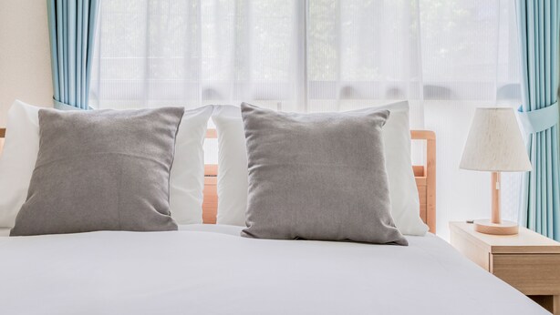 Una cama bien hecha con dos almohadas grises, una colcha blanca y una mesita de noche de madera con una lámpara.